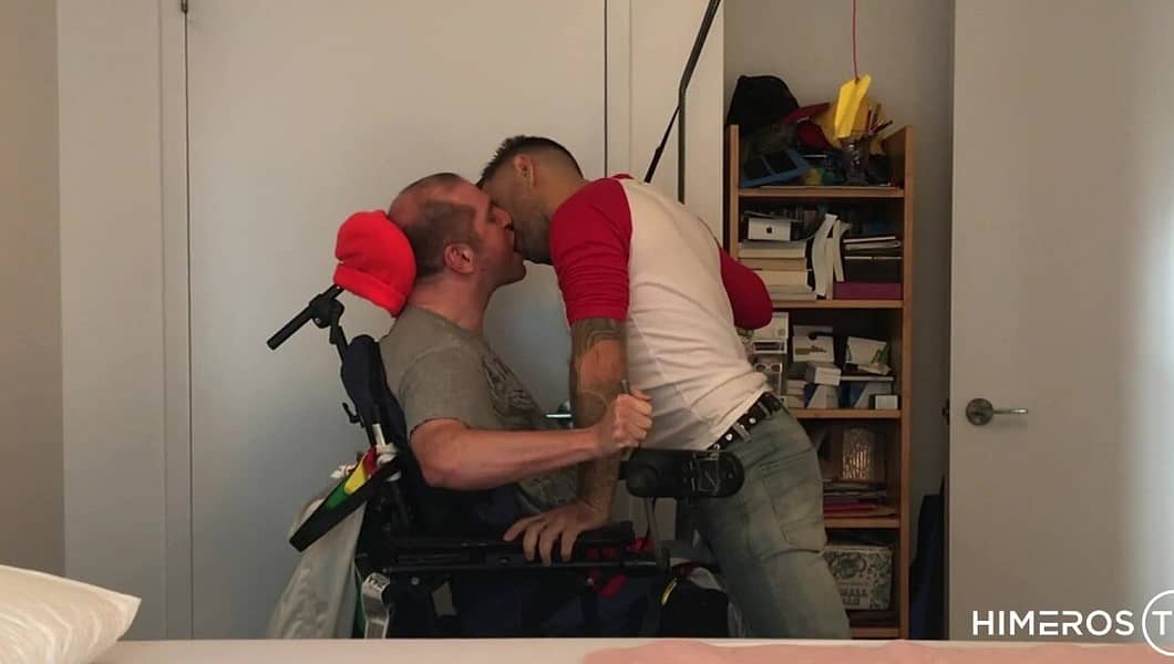 A white disabled man in a wheelchair is kissed by a standing man leaning into him.