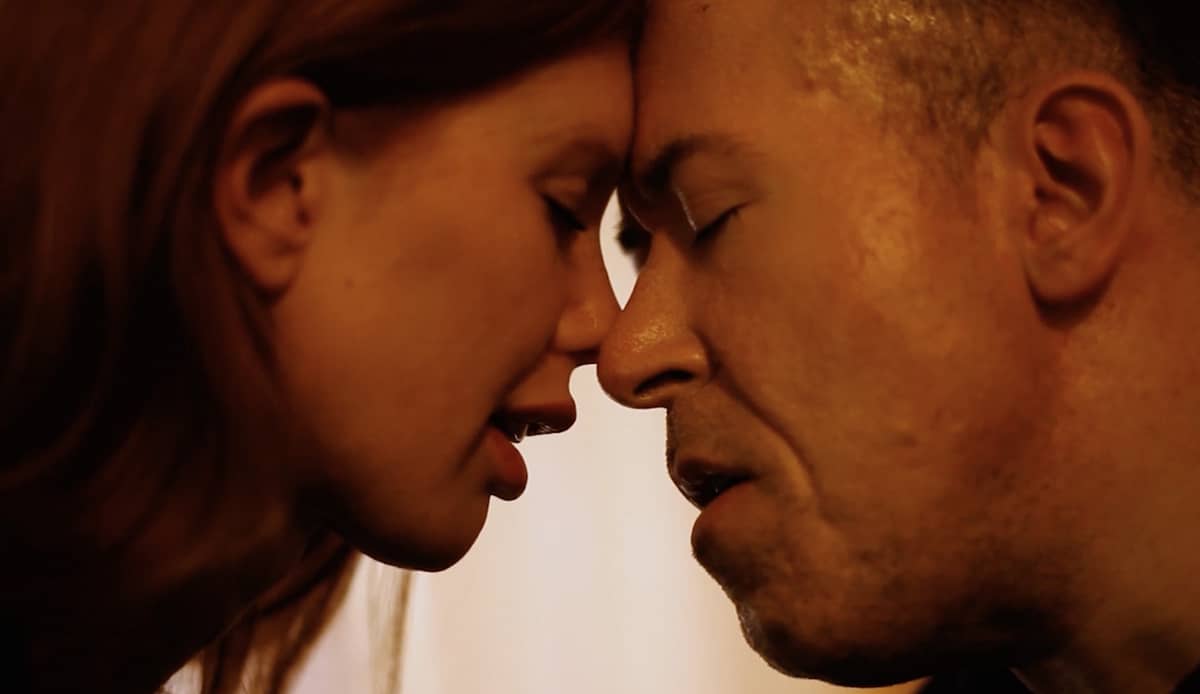 A still from the adult film Into-You-I-See-Me shows a close up of disabled porn performer Oliver David and performer Rachel Rose with their eyes closed, intimately leaning their faces into each other.