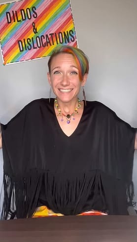 A white woman in black sits in a wheelchair. Short rainbow hair matches rainbow sign "Dildos and Dislocations."