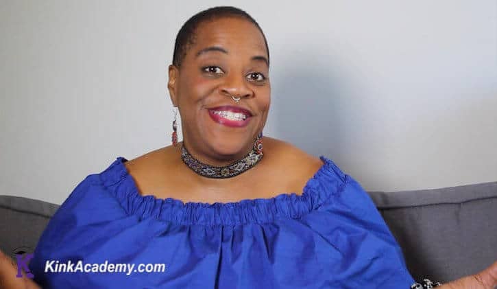 Robin Wilson-Beattie, a woman of color, smiles while wearing a blue top. She appears in a screenshot for a virtual course on BDSM with a disability for KinkAcademy.Com.